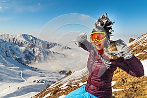 Emotional traveler young woman with windy hair smiling, sitting on top of sunny mountains. Funny woman in mohawk hat