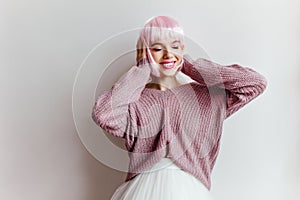 Emotional trendy girl with shiny pink hair standing in front of white wall. Indoor photo of carefree lady in peruke and