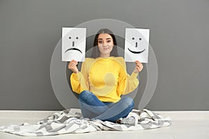 Emotional woman holding sheets of paper with drawn emoticons while sitting near grey wall