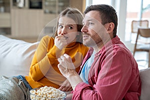 Emotional woman and intrigued man watching thriller, action film, scary movie at home eating popcorn