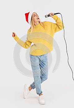 Emotional woman in Santa Claus hat singing with microphone on light grey background. Christmas music