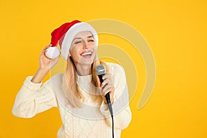 Emotional woman in Santa Claus hat singing with microphone on yellow background. Christmas music
