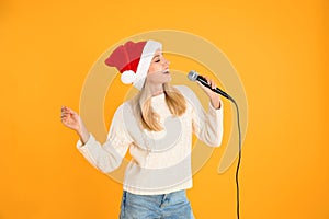 Emotional woman in Santa Claus hat singing with microphone on yellow. Christmas music