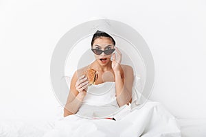 Emotional woman with towel on head lies in bed under blanket isolated over white wall background wearing sunglasses eat burger