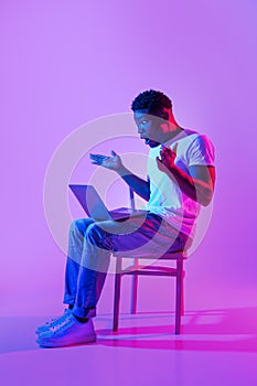 Emotional young black man sitting on chair with laptop, feeling shocked, making mistake in business project in neon