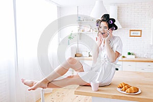 Emotional young careless woman sit on table and talk on phone. Amazed housekeeper in kitchen. Wearing white dressing