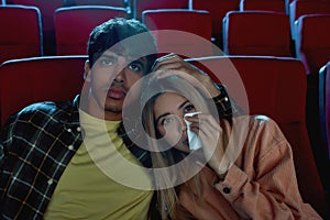Emotional young couple, attractive man and woman sitting at the cinema, watching sad movie together. Girlfriend wiping