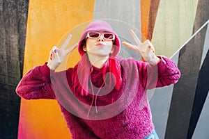 Emotional young hipster woman with pink hair and sunglasses in magenta fluffy sweatshirt and bucket hat making V sign by