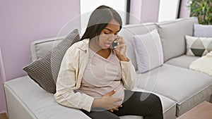 Emotional young latina expecting mother, casually touching belly on living room sofa, has a serious talk on her smartphone as