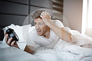 Emotional young man on bed in early morning. He overslept. Guy hold clock and look at it scared.