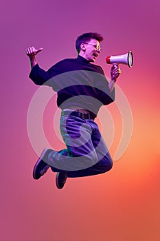 Emotional young man in casual clothes jumping and talking megaphone against gradient pink background in neon light