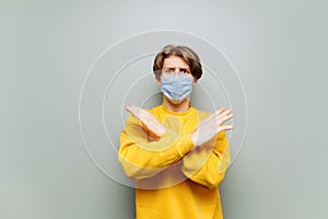 Emotional young man with crossed arms stands on a gray background with a medical protective mask on his face and stares blankly at