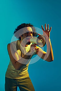 Emotional young man with curly hair shouting in megaphone against blue background in neon light