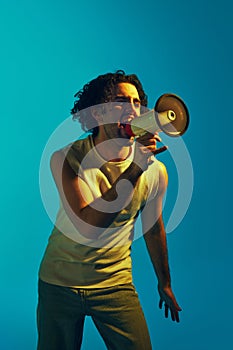 Emotional young man with curly hair shouting in megaphone against blue background in neon light. News, propaganda