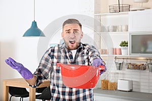 Emotional young man holding plastic basin under water leakage from ceiling in kitchen.