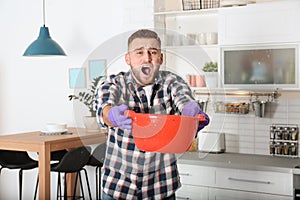 Emotional young man holding plastic basin under water leakage from ceiling in kitchen.