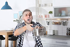 Emotional young man holding saucepan under water leakage from ceiling in kitchen, space for text