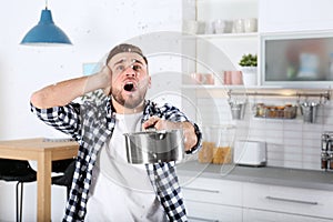 Emotional young man holding saucepan under water leakage from ceiling in kitchen, space for text.