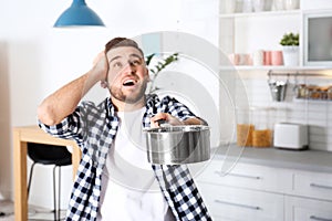 Emotional young man holding saucepan under water leakage from ceiling in kitchen, space for text