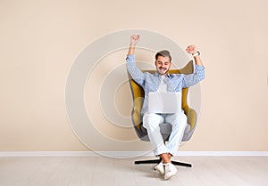 Emotional young man with laptop in armchair near color wall indoors