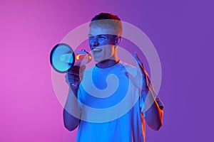 Emotional young man shouting in megaphone against gradient pink purple background in neon light. News, sales