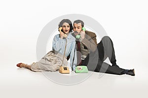 Emotional young man and woman sitting on floor and talking on vintage rotary phone isolated on white background