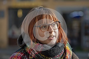 An emotional young woman of 20-25 years old with glasses, dyed red hair and a little overweight against the background of the city