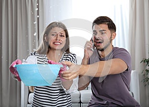 Emotional young woman collecting water leaking from ceiling while her husband calling plumber