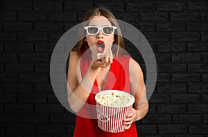 Emotional young woman with cup of popcorn and 3D cinema glasses against dark brick wall
