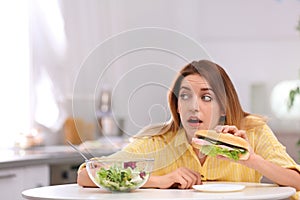 Emotional young woman eating sandwich instead of salad in kitchen