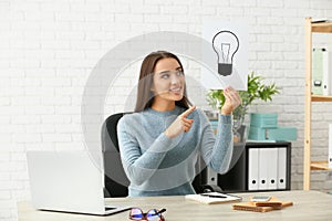 Emotional young woman holding paper sheet with drawn light bulb as symbol of idea while sitting at table