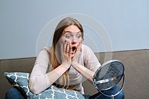 Emotional young woman looking at make-up mirror while sitting at home on bed