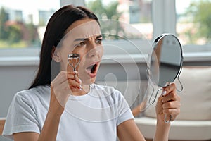 Emotional young woman with mirror using eyelash curler indoors