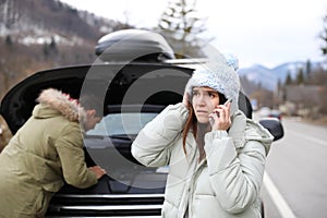 Emotional young woman talking on phone while stressed man repairing broken car in winter