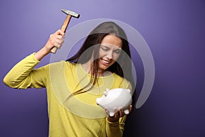 Emotional young woman trying to break a piggy bank with hammer on color background