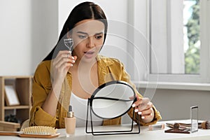 Emotional young woman using eyelash curler while doing makeup at table indoors