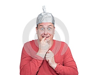 Emotionally scared man wearing aluminum foil hat holds finger in mouth, isolated on white background.