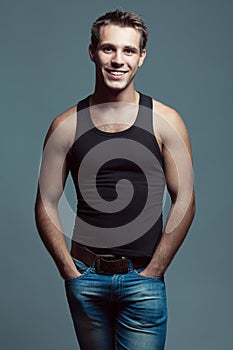 Emotive portrait of handsome young man looking at camera with arms in pockets of blue jeans. White shiny smile, great muscles and