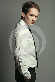 Emotive portrait of serious handsome young man in casual white shirt, black tie, leather belt, gray pants posing over grey