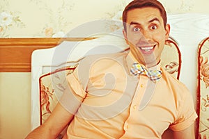 Emotive portrait of smiling handsome crazy hipster in trendy casual shirt and colorful bow-tie sitting on a white vintage sofa in