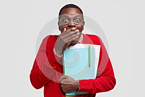 Emotive surprised dark skinned young man covers mouth with palm, has joyful expression, recieves excellent mark on exam