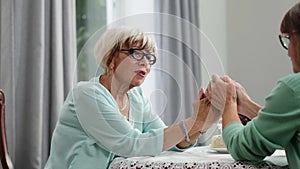 Empathic senior blond woman in eyeglasses holding hands with friend and talking. Portrait of confident Caucasian retiree