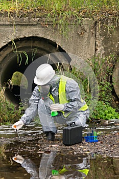An employee in a protective suit and mask takes water from the river with a pipette and conducts a test of the suitability of photo