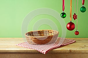 Empty basket on wooden table with hanging ornament decoration and tablecloth over green background. Christmas mock up for design