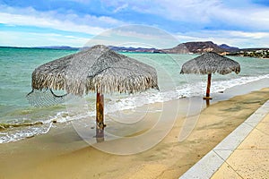 Empty beach with two palapas next to promenade, sea and mountains in background