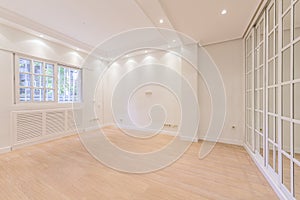 Empty bedroom with white painted walls, light wooden floor, French-style aluminum windows, radiator covers of the same material