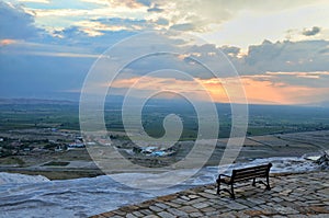 An empty bench in Pamukkale, Turkey, facing the immensity of the horizon, houses, hotels, green fields, and a sunset coloring the