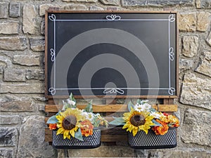 an empty black blackboard hanging on a stone wall with some decorative flowers below it, copy space