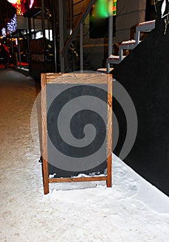 Empty black board with a wooden frame and colorful lights, on the snow around the street, winter and evening city