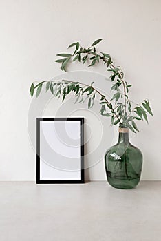 Empty black picture frame mockup and green glass jar with tree branches on neutral beige table, concrete wall background